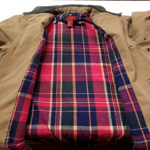 May include: A tan work jacket with a dark brown collar. The interior lining features a red, blue, and green plaid pattern. The jacket has a button closure and a classic design.