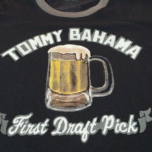 May include: A black t-shirt featuring a graphic of a beer mug filled with golden liquid and white foam. The text TOMMY BAHAMA is printed in white above the mug, and the text First Draft Pick is printed in white below the mug on a banner design.