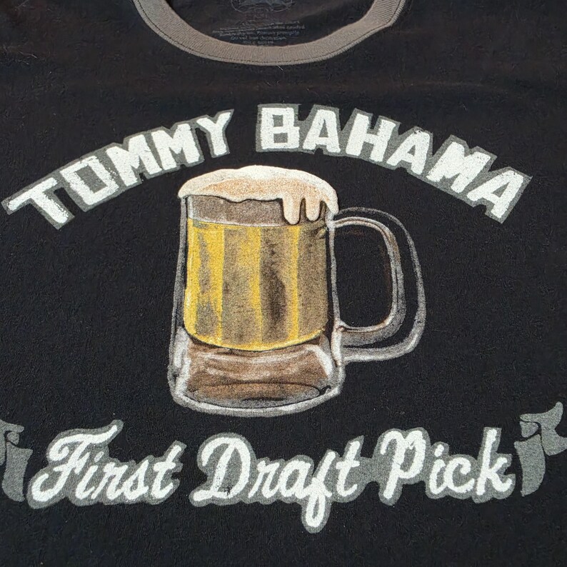 May include: A black t-shirt featuring a graphic of a beer mug filled with golden liquid and white foam. The text TOMMY BAHAMA is printed in white above the mug, and the text First Draft Pick is printed in white below the mug on a banner design.