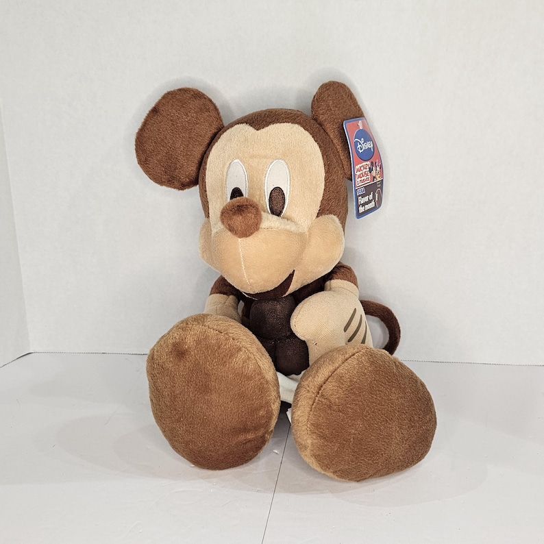 Puede incluir: Un peluche de Mickey Mouse en marr&oacute;n, beige y blanco. El peluche tiene grandes orejas marrones, una cara beige y pies marrones. Tiene una etiqueta que dice "Disney".
