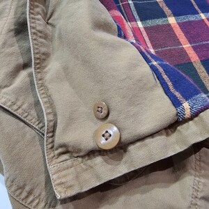 May include: Detail of a tan jacket with two brown buttons. The jacket features a plaid lining in blue, red, and purple. The fabric looks like a sturdy cotton or canvas material.