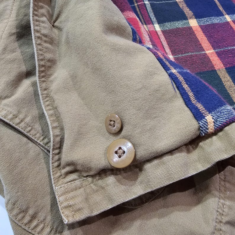 May include: Detail of a tan jacket with two brown buttons. The jacket features a plaid lining in blue, red, and purple. The fabric looks like a sturdy cotton or canvas material.