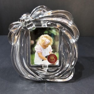 May include: Clear glass picture frame with a bow-shaped design. The frame holds a photograph of a young child in a pink dress. The frame has a decorative, swirling pattern and a small, round sticker.
