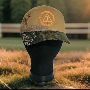 May include: A tan baseball cap with a camouflage brim. The cap features the text "TRIANGLE CONSTRUCTION SERVICES" in orange, along with a deer silhouette. The cap is displayed on a black mannequin head.