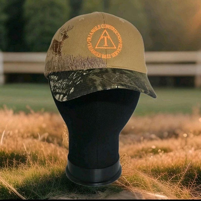 May include: A tan baseball cap with a camouflage brim. The cap features the text "TRIANGLE CONSTRUCTION SERVICES" in orange, along with a deer silhouette. The cap is displayed on a black mannequin head.