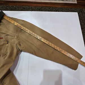 May include: A tan work jacket with a dark green collar and a small chest pocket. A wooden ruler is placed on the jacket, measuring its length. The text "BLOUNT COUNTY COOPERATIVE, INC." is visible on the ruler.