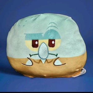 May include: A plush, round pillow featuring the face of Squidward Tentacles from the animated series Spongebob Squarepants. The pillow has a light blue top, a tan bottom, and Squidward's face with brown eyes and a turquoise nose.