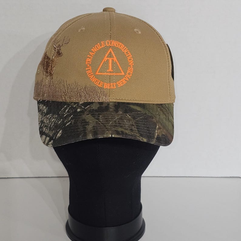 May include: Tan baseball cap with a camouflage brim. The cap features an embroidered deer and the words "TRIANGLE CONSTRUCTION TRIANGLE BELT SERVICES" in orange. A black tag is visible on the side.