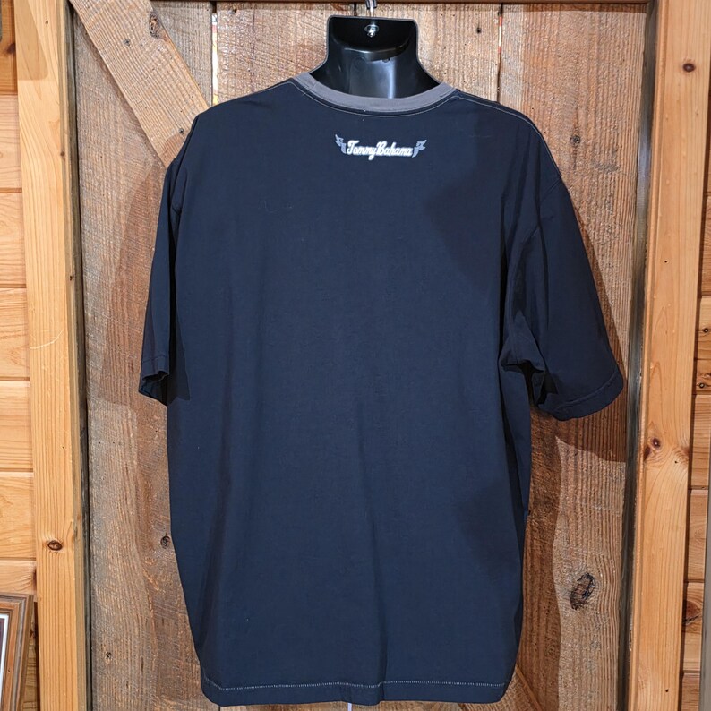 May include: A short-sleeved black t-shirt featuring a grey crew neckline and the white text Tommy Bahama printed on the upper back area. The garment is displayed on a black mannequin torso, showcasing a solid black fabric construction throughout.
