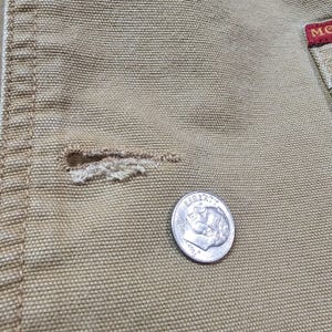 May include: Close-up of tan fabric with a buttonhole and a dime. The buttonhole is frayed. The dime shows the profile of a person and the word "LIBERTY". The fabric has a textured weave.