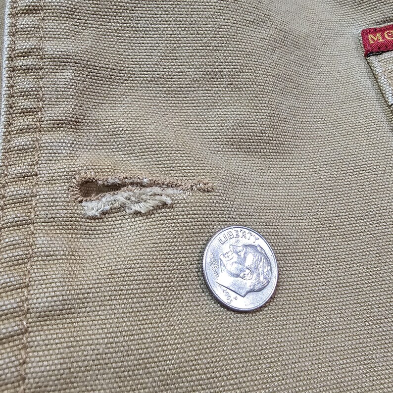 May include: Close-up of tan fabric with a buttonhole and a dime. The buttonhole is frayed. The dime shows the profile of a person and the word "LIBERTY". The fabric has a textured weave.
