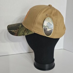 May include: A tan baseball cap with a camouflage brim. The cap has an orange logo on the side and a hang tag with a landscape design. The cap is displayed on a black mannequin head.