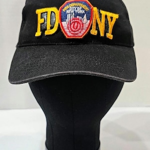 May include: Black baseball cap with the text "FDNY" in yellow block letters. The cap features a patch with the words "Fire Department City of New York" and a cityscape design. The cap is displayed on a black mannequin head.