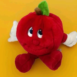 May include: A plush red apple character with a smiling face, white gloves, and white wings. The apple has a green leaf and a brown stem. The toy is on a yellow background.