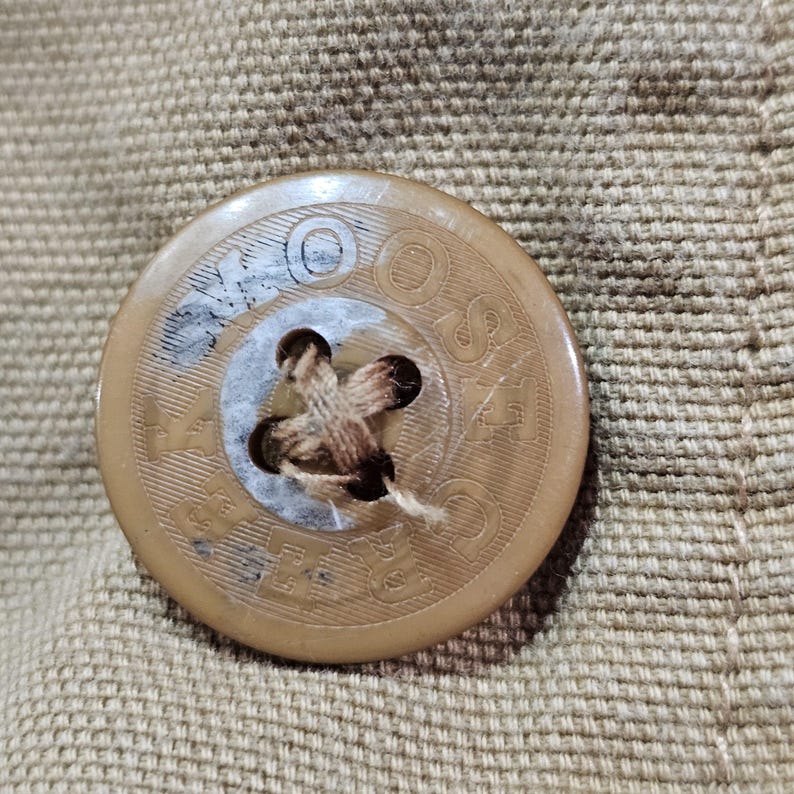 May include: Close-up of a tan button with the word "SASCO" and other text around the edge. The button has four holes and is attached to tan fabric.