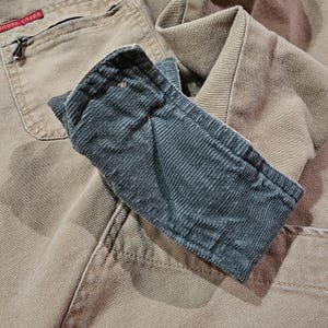 May include: A close-up shot of a tan work jacket with a dark grey corduroy sleeve. The jacket features a zipped pocket and a red tag with the brand name "Goose Creek". The corduroy sleeve is folded over the jacket.