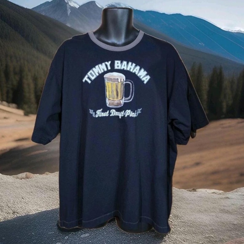 May include: A black short-sleeve t-shirt with a grey crew neckline. The front features a graphic of a beer mug with foam, the text "TOMMY BAHAMA" arched above, and the text "First Draft Pick" on a banner below the mug in white lettering.