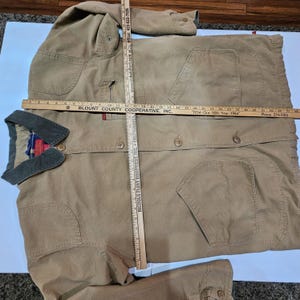May include: A tan work jacket with a green collar and multiple pockets. The jacket has button closures and a plaid pattern on the inside collar. Measurements are being taken with a ruler.
