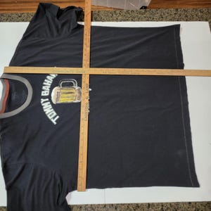 May include: A dark gray short-sleeved t-shirt with a gray crew neckline. The shirt features a graphic of a beer mug and the text Tommy Bahama. Two wooden rulers are placed over the shirt, showing a width of 26 inches and a length of 28 inches.