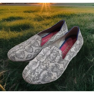 May include: A pair of slip-on shoes featuring a grey and white snakeskin pattern knit fabric. The interior of the shoes is lined with a bright pink insole that displays the brand name Rothy's in white text, with a blue trim along the collar.