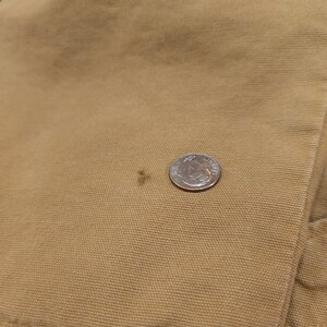 May include: Close-up of tan fabric with a small dark stain and a US dime coin for scale. The fabric appears to be a durable material, possibly canvas or a similar workwear fabric. The dime is approximately 1.8 cm in diameter.