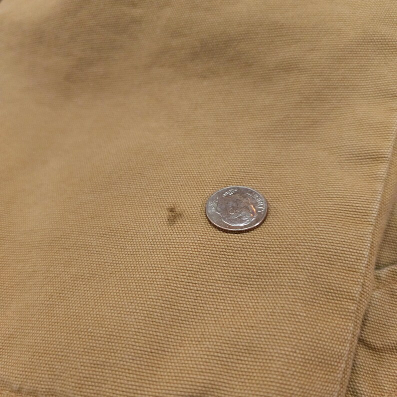 May include: Close-up of tan fabric with a small dark stain and a US dime coin for scale. The fabric appears to be a durable material, possibly canvas or a similar workwear fabric. The dime is approximately 1.8 cm in diameter.