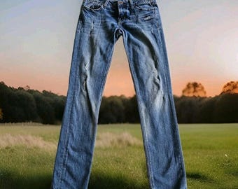 May include: Faded blue denim jeans with distressed details, including rips on the knees. The jeans have a classic five-pocket design and a button and zipper closure. The jeans are displayed against a backdrop of a field and trees.