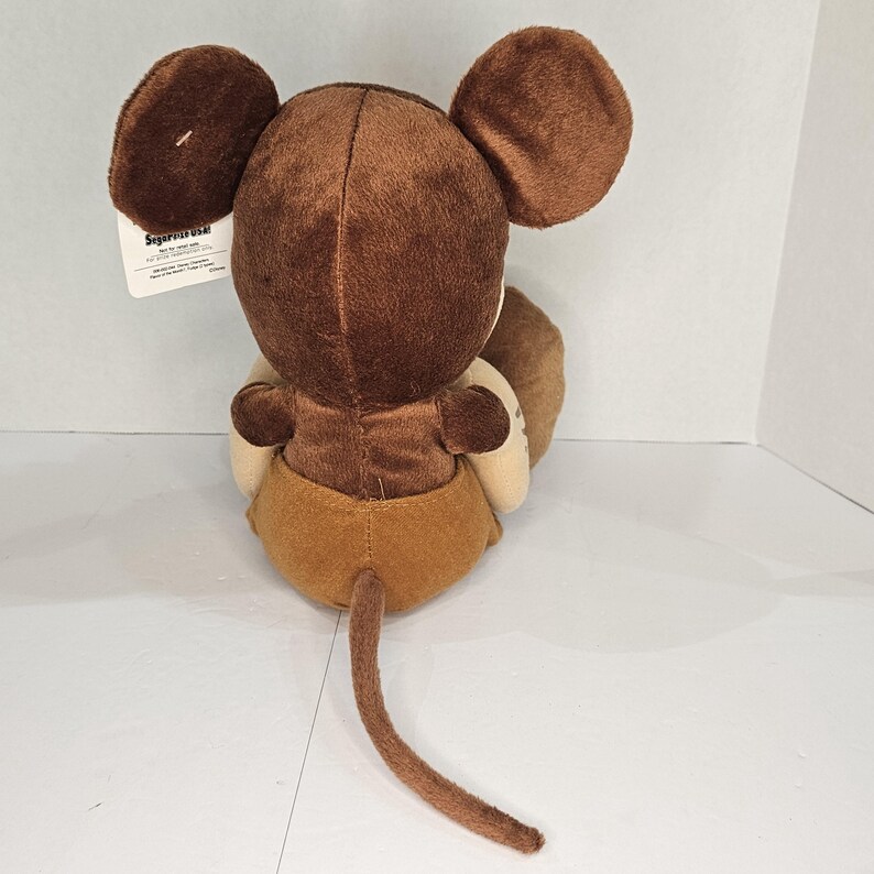 Puede incluir: Un peluche marr&oacute;n con forma de rat&oacute;n con orejas grandes, una cola larga y una barriga beige. El juguete est&aacute; sentado, de espaldas al espectador. Una etiqueta est&aacute; adherida al juguete.