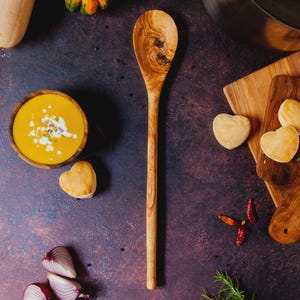 May include: A wooden spoon with a long handle and a rounded head rests on a dark, textured surface. A bowl of orange soup, heart-shaped biscuits, and red onions are also visible, suggesting a culinary theme.