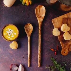 May include: Overhead shot of a culinary scene featuring a wooden bowl of orange soup, two wooden spoons, heart-shaped pastries, and a wooden cutting board. The scene includes red onions, chili peppers, and a dark purple surface.