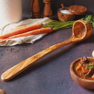 May include: An olive wood soup ladle with a long handle and a large, rounded bowl. The ladle displays a natural wood grain. The background features carrots, a bowl of soup, and other kitchen items.