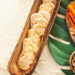 May include: A wooden serving tray filled with small, round, lightly browned crackers. The tray is long and narrow, with a natural wood finish. The crackers are arranged in a single row. Other food items are visible in the background.