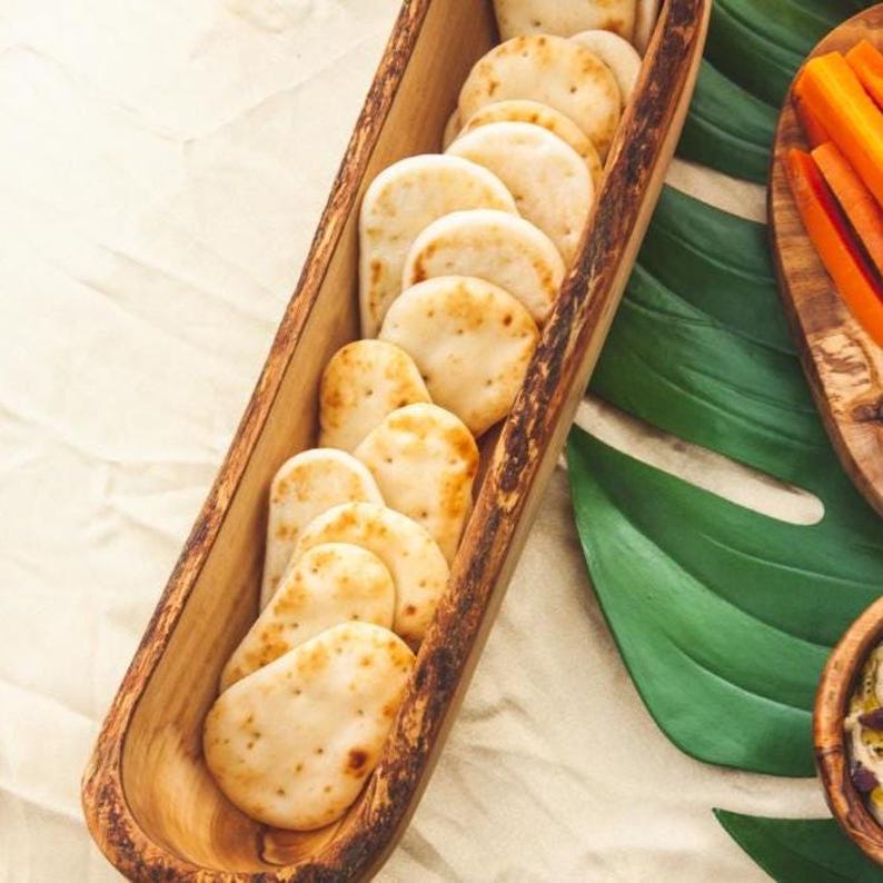 May include: A wooden serving tray filled with small, round, lightly browned crackers. The tray is long and narrow, with a natural wood finish. The crackers are arranged in a single row. Other food items are visible in the background.