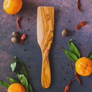 Può includere: Una spatola di legno con un motivo a venature marrone chiaro, circondata da arance, peperoncini rossi, noce moscata e foglie verdi. La spatola è in legno d'ulivo ed è un utensile da cucina.
