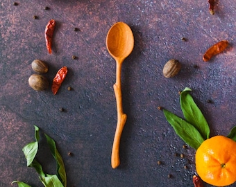 Puede incluir: Una cuchara de madera de color marrón claro con un mango con forma de rama natural. La cuchara está rodeada de cítricos naranjas, hojas verdes, chiles rojos secos, nueces marrones y granos de pimienta negra sobre una superficie oscura.