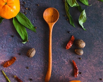 Puede incluir: Una cuchara de madera con un mango largo y delgado y un cuenco redondo, colocada verticalmente. La cuchara está rodeada por dos cítricos de color naranja brillante con hojas verdes, varios chiles rojos secos y pequeñas nueces moscadas.