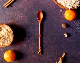 Puede incluir: Una cuchara de madera de mango largo con un cuenco redondeado y liso y un detalle decorativo trenzado en la base del mango, colocada verticalmente en el centro, rodeada de ramas de canela, frutas naranjas y cuencos con avena y azúcar.