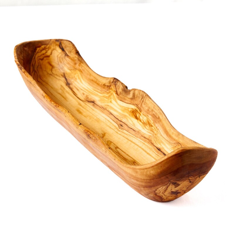 May include: A handcrafted, wooden serving bowl with a natural, organic shape. The bowl is made from olive wood, showcasing a warm, light brown color with unique grain patterns. The elongated design is ideal for serving snacks or displaying decorative items.