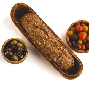 May include: A wooden serving set featuring a long, oval-shaped bread bowl with a seeded baguette. Two small wooden bowls contain olives and colorful cherry tomatoes. The set is arranged on a white background.