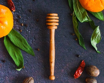 Puede incluir: Un cucharón de miel de madera con un mango largo y delgado y una cabeza ranurada, colocado verticalmente. El cucharón está rodeado por dos cítricos de color naranja brillante con hojas verdes, varios chiles rojos secos y nueces marrones.
