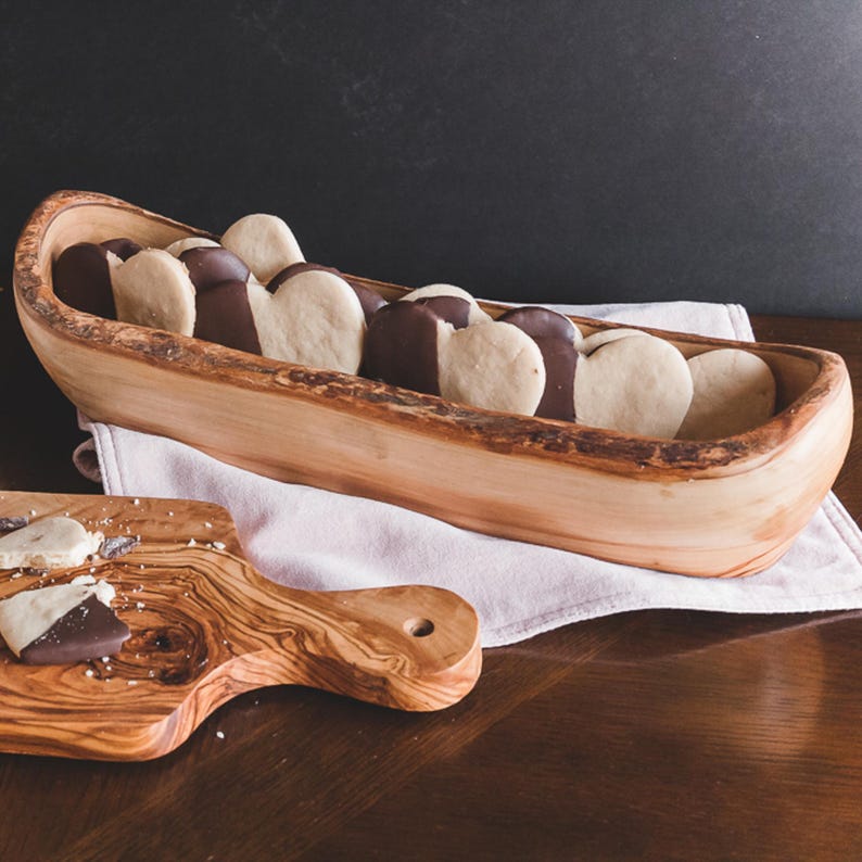 May include: A wooden serving bowl filled with heart-shaped cookies dipped in chocolate. The cookies rest on a white cloth inside the bowl. A wooden cutting board with cookie crumbs is in the foreground.