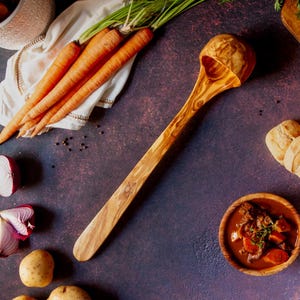 May include: A wooden cooking spoon with a rounded bowl and long handle. The spoon is made of light-colored wood with a natural grain pattern. The image also includes carrots, potatoes, red onion, and a bowl of stew.