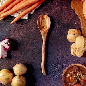 Puede incluir: Una cuchara de madera con un mango largo descansa sobre una superficie oscura y texturizada, rodeada de ingredientes. Zanahorias frescas, patatas y un cuenco de estofado son visibles. La escena sugiere una comida rústica y casera.