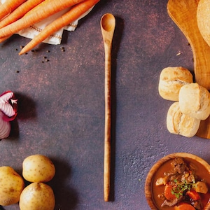 Könnte beinhalten: Ein langer Holzlöffel mit abgerundeter Schüssel und schlankem Griff. Der Löffel ist von Zutaten für eine Mahlzeit umgeben, darunter Karotten, Kartoffeln und eine Schüssel Eintopf. Der Hintergrund ist eine dunkle, strukturierte Oberfläche.