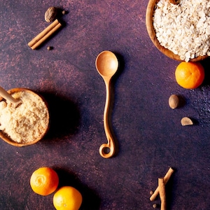 May include: A light brown wooden spoon with a long, slender, curved handle that ends in a small circular loop. The spoon is positioned vertically between two wooden bowls, one containing brown sugar and the other filled with rolled oats.