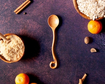 Puede incluir: Una cuchara de madera de color marrón claro con un mango largo, delgado y curvado que termina en un pequeño bucle circular. La cuchara está colocada verticalmente entre dos cuencos de madera, uno con azúcar moreno y otro con avena.