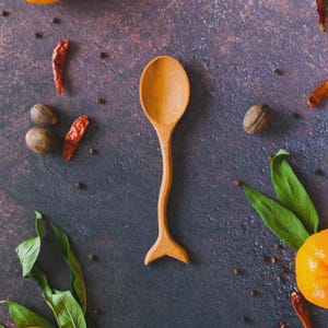 Puede incluir: Una cuchara de madera con un mango curvo único y un cuenco en forma de cuchara. La cuchara está rodeada de especias, incluyendo chiles rojos, granos de pimienta y nuez moscada. Hojas verdes y una mandarina añaden color a la composición.