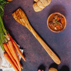 Könnte beinhalten: Ein Holzspatel mit breitem, flachem Kopf und langem Griff, neben frischen Karotten, einer Schüssel Eintopf und Brötchen. Der Spatel ist aus hellem Holz mit natürlicher Maserung gefertigt. Der Eintopf enthält Fleisch- und Gemüsestücke.