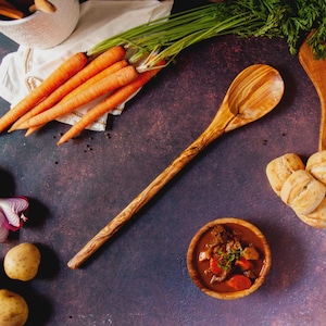 May include: A wooden spoon with a long handle lies on a dark, textured surface, surrounded by fresh ingredients. Bright orange carrots, a bowl of stew, and small rolls are arranged around the spoon, suggesting a rustic, home-cooked meal.