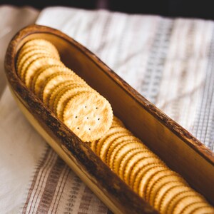 May include: A wooden serving dish filled with round, golden-brown crackers. The crackers are arranged in a row inside the dark wooden dish. The dish sits on a white and brown striped cloth.